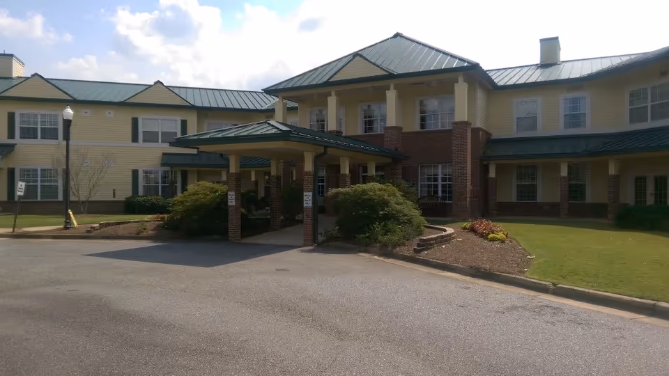 Front entrance of a two-story senior living building with a covered porte-cochere, yellow siding, brick accents, and a green metal roof.