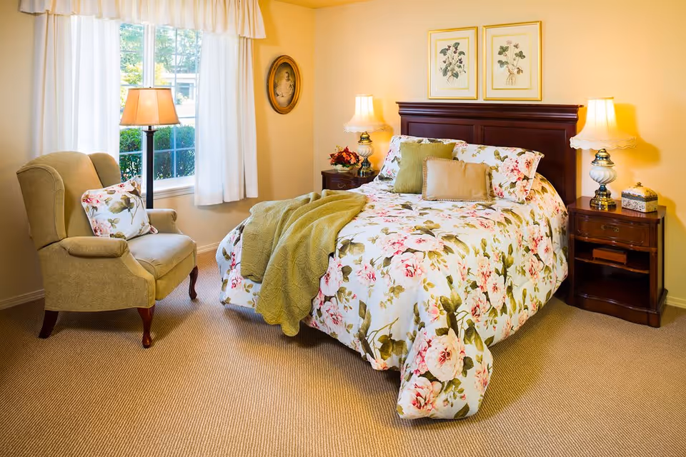 A cozy bedroom with a floral-patterned bedspread on a wooden bed frame, two bedside tables each with a lamp, a green armchair with a floral pillow, a window with white curtains, and framed botanical prints on the wall.