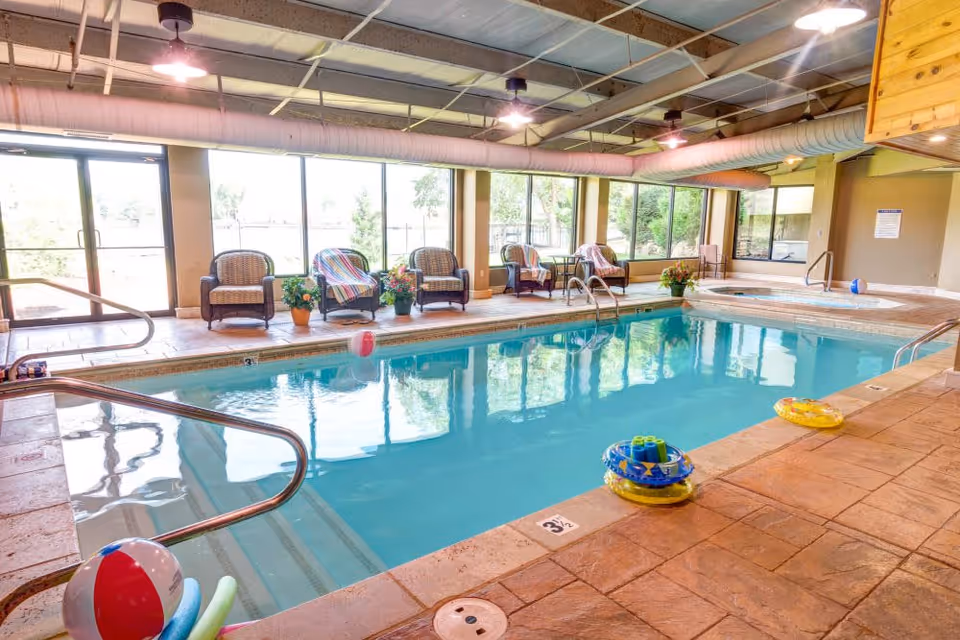 Indoor swimming pool area with clear blue water, surrounded by a tiled floor. Several wicker chairs with colorful blankets and potted plants are arranged along the large windows that let in natural light. Pool toys and floatation devices are visible in and around the pool. The ceiling has exposed ducts and lighting fixtures.