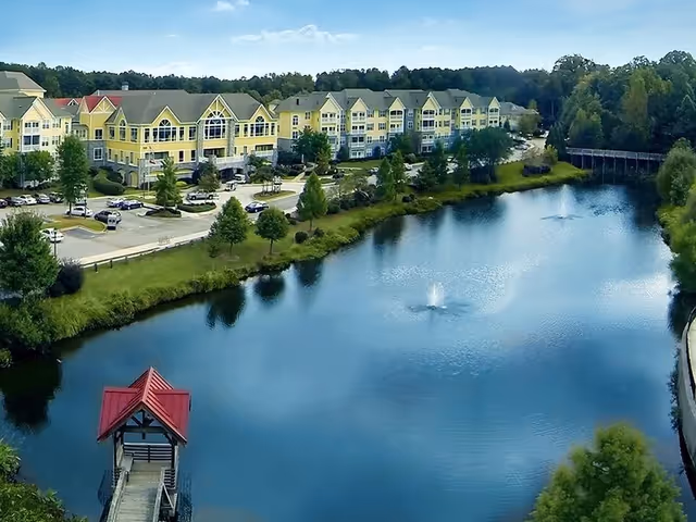 Aerial view of a yellow senior living complex beside a pond with fountains and a small red-roofed gazebo.