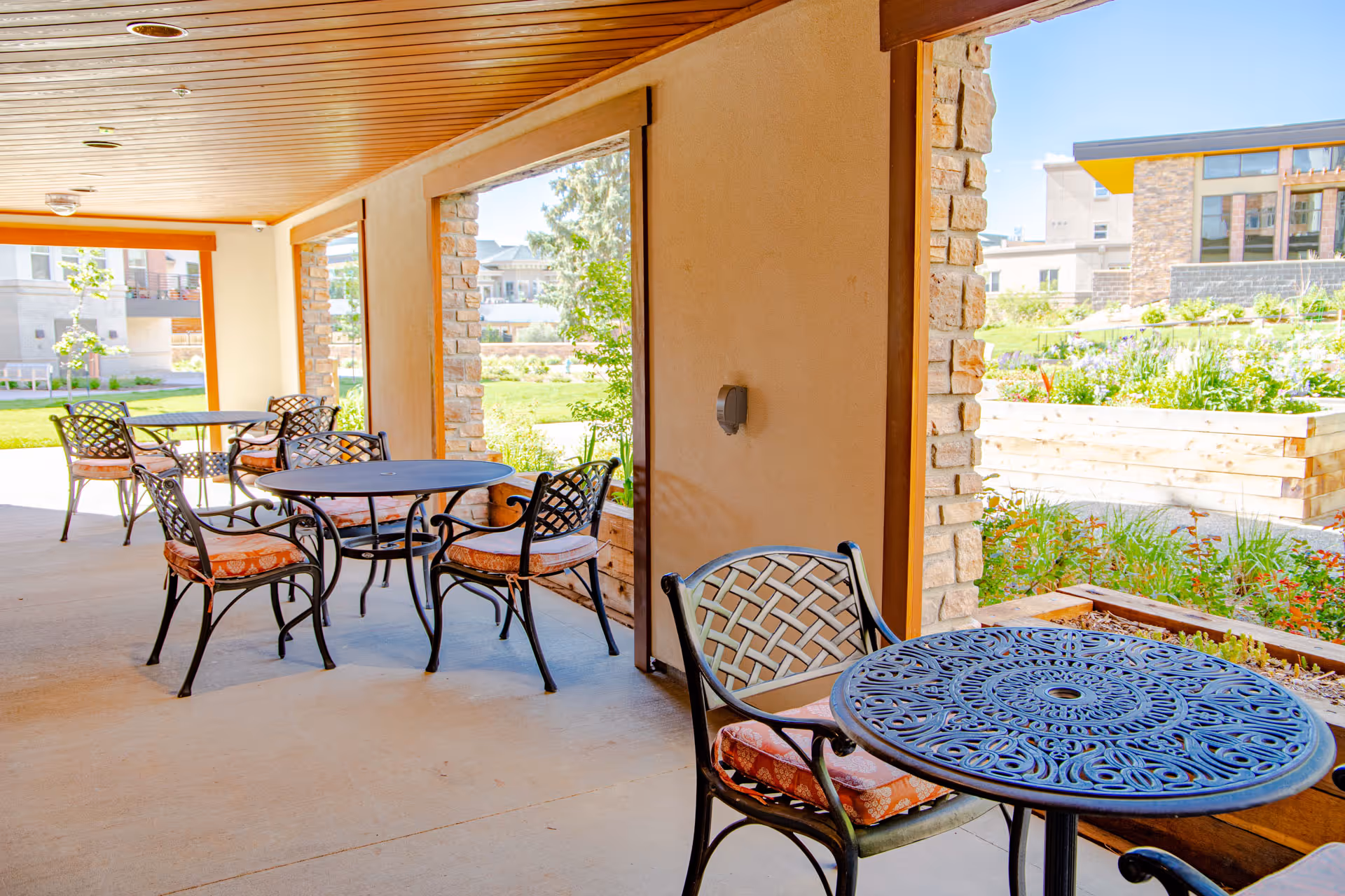 Covered outdoor patio area with several round metal tables and cushioned chairs. The patio overlooks a garden with raised wooden planters and greenery, with buildings visible in the background.