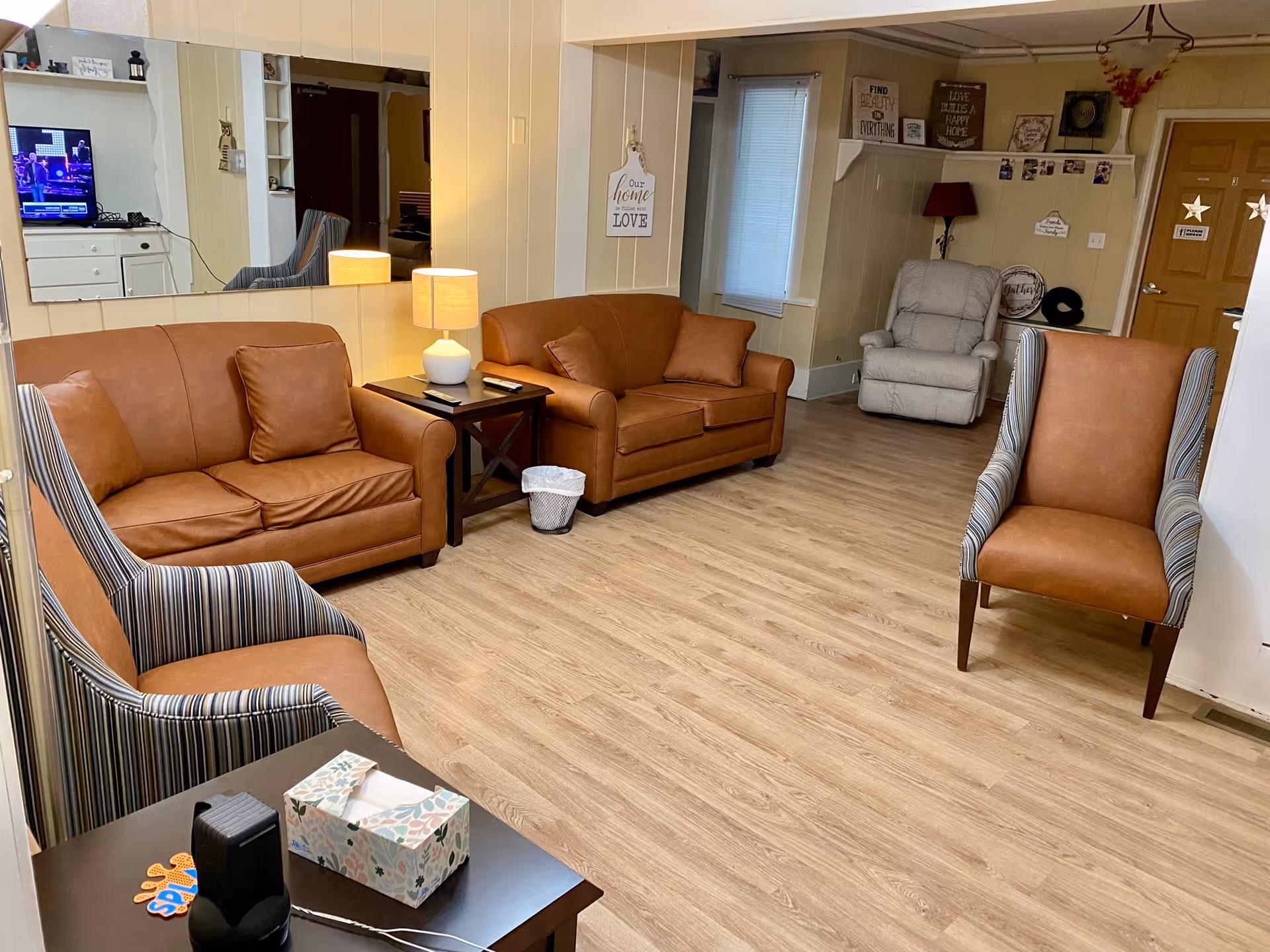 A cozy communal living room with leather sofas, striped armchairs, a side table with lamps, and wood-look flooring.