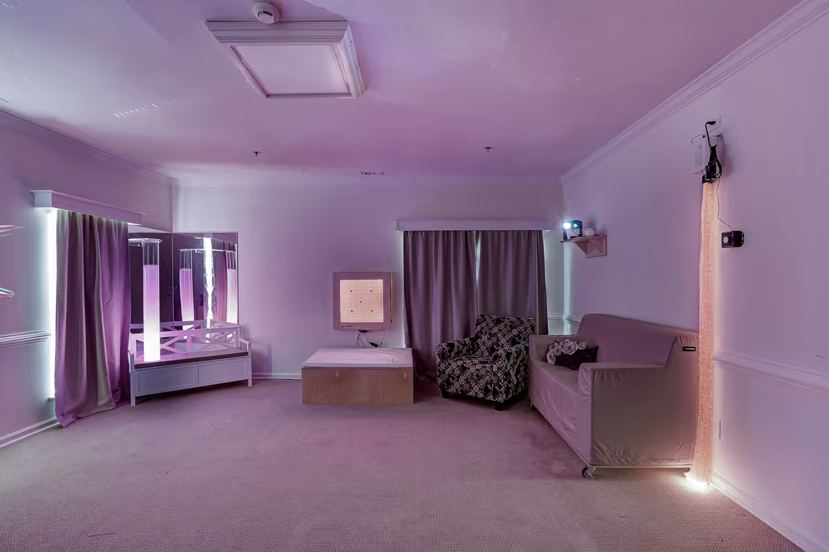 A softly lit room with purple lighting featuring a patterned armchair, a covered sofa, a low wooden platform, and a mirrored vanity with vertical light tubes. The room has carpeted flooring, curtains covering windows, and some mounted electronic devices on the walls.