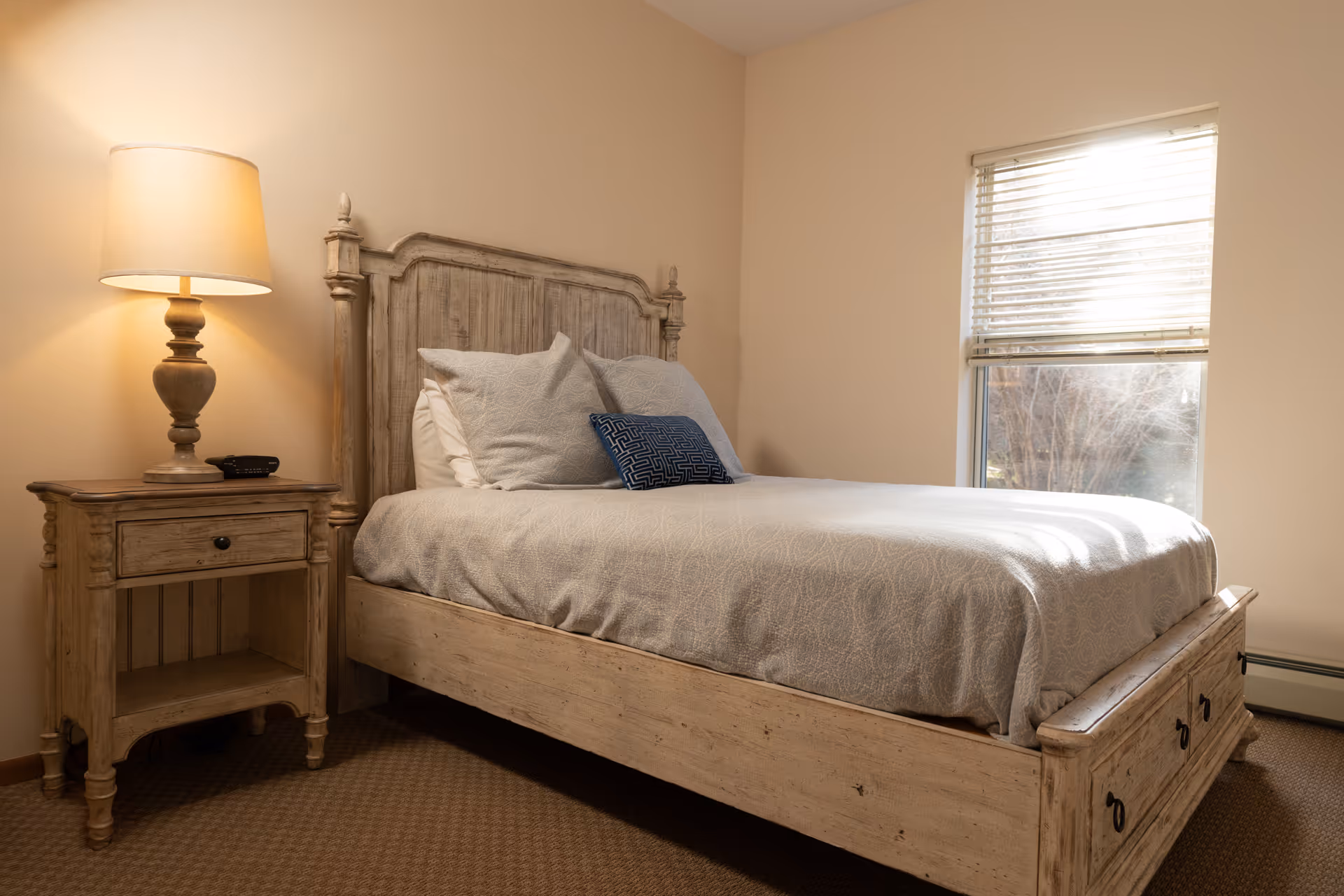 A cozy bedroom with a wooden bed, bedside table and lamp next to a window.