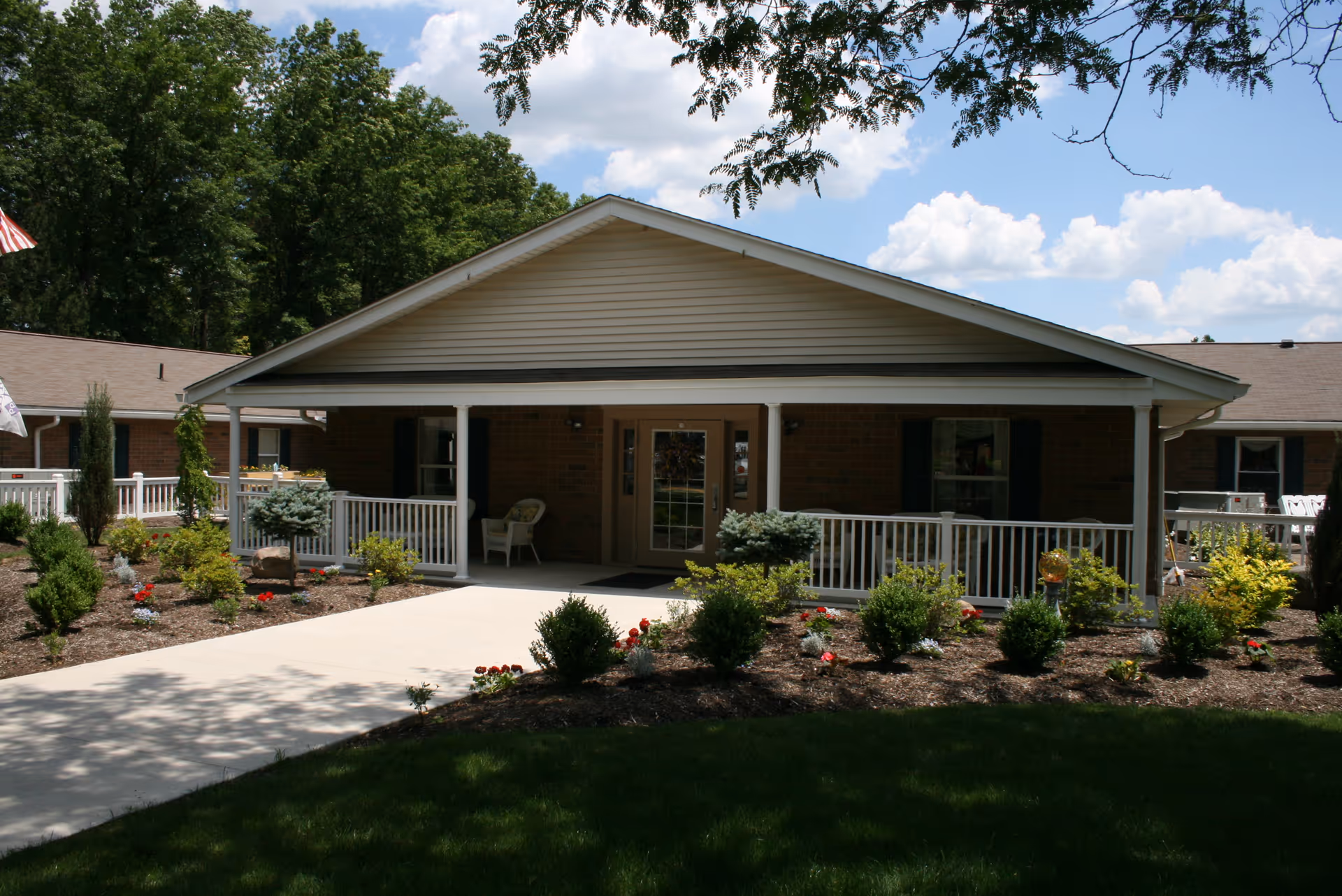 Front exterior view of a single-story brick building with a covered porch, white railings, and outdoor chairs. The building is surrounded by landscaped garden beds with shrubs and flowers, and there are trees and a partly cloudy sky in the background.
