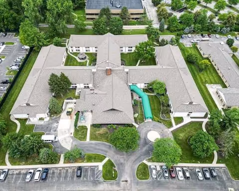 Aerial view of a U-shaped senior living facility building with landscaped lawns, trees, walkways, and parked cars.