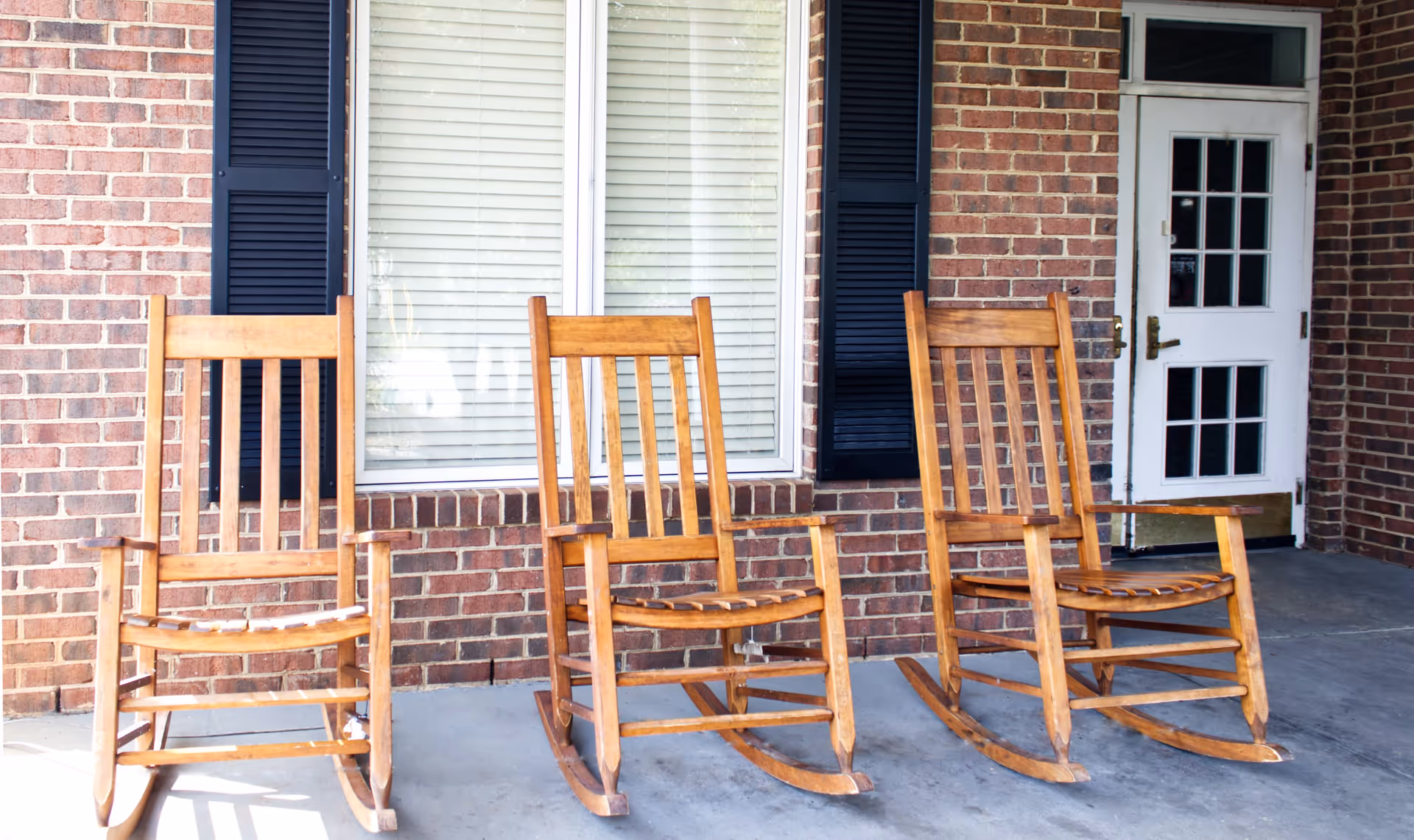 Three wooden rocking chairs placed on a concrete porch in front of a brick wall with a window and a white door with glass panes.