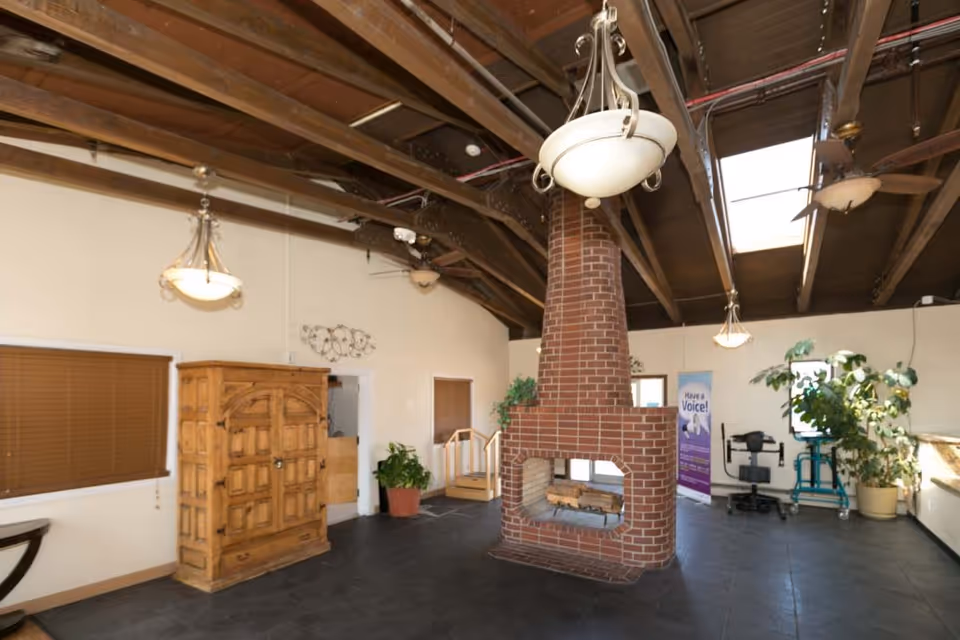A spacious room with a high wooden ceiling featuring exposed beams and skylights. In the center is a large brick fireplace with a chimney extending to the ceiling. The room has several hanging light fixtures and ceiling fans. There are wooden blinds on the windows, a wooden cabinet, potted plants, and some medical equipment near the back wall. A banner with the text 'Have a Voice!' is visible in the background.