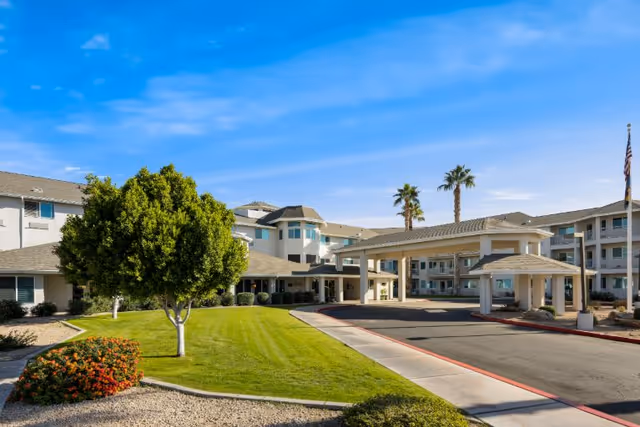 Exterior view of Vista del Rio - A Provincial Senior Living Community, showing a large building with multiple floors, a covered entrance driveway, well-maintained green lawn, trees, and clear blue sky.