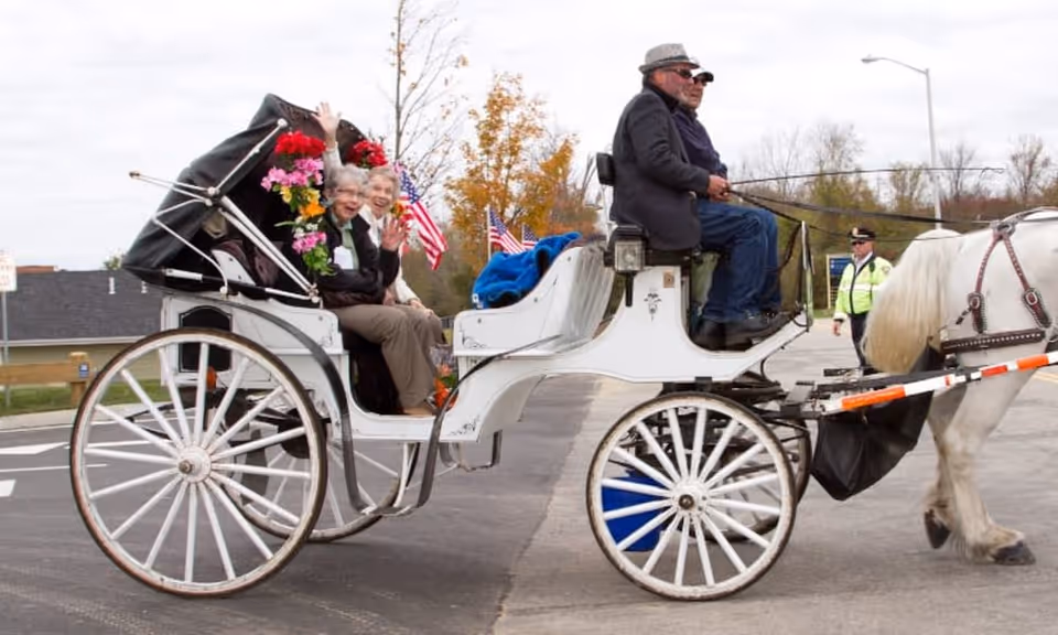 Two people wave from a white horse-drawn carriage driven by a man on a paved street with the horse partly visible.