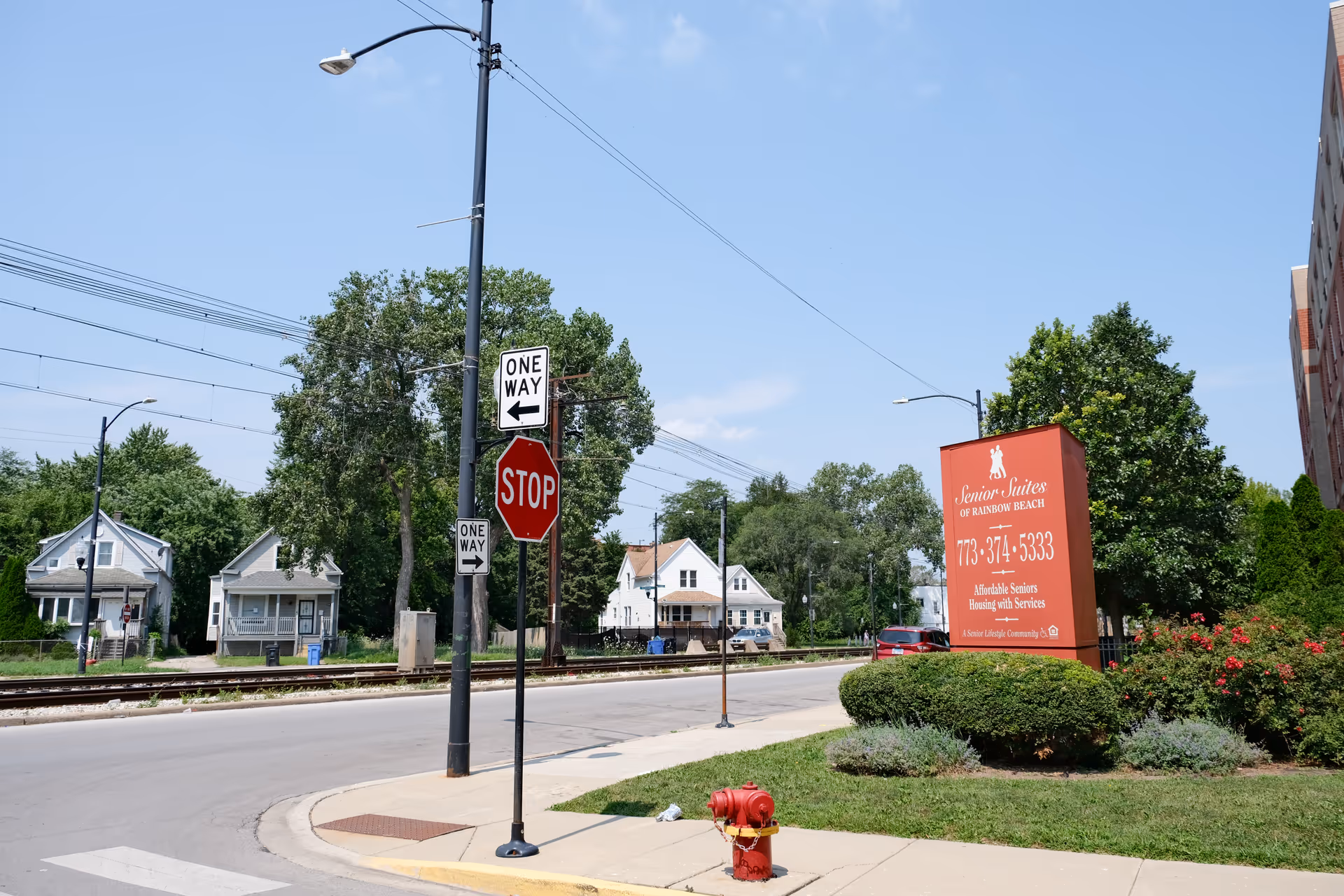 Street view showing a stop sign and one way signs near a railroad track. On the right side, there is a red sign for Senior Suites of Rainbow Beach with contact information and a description of affordable seniors housing with services. Surrounding the sign are bushes and flowers, and there are houses and trees in the background under a clear blue sky.
