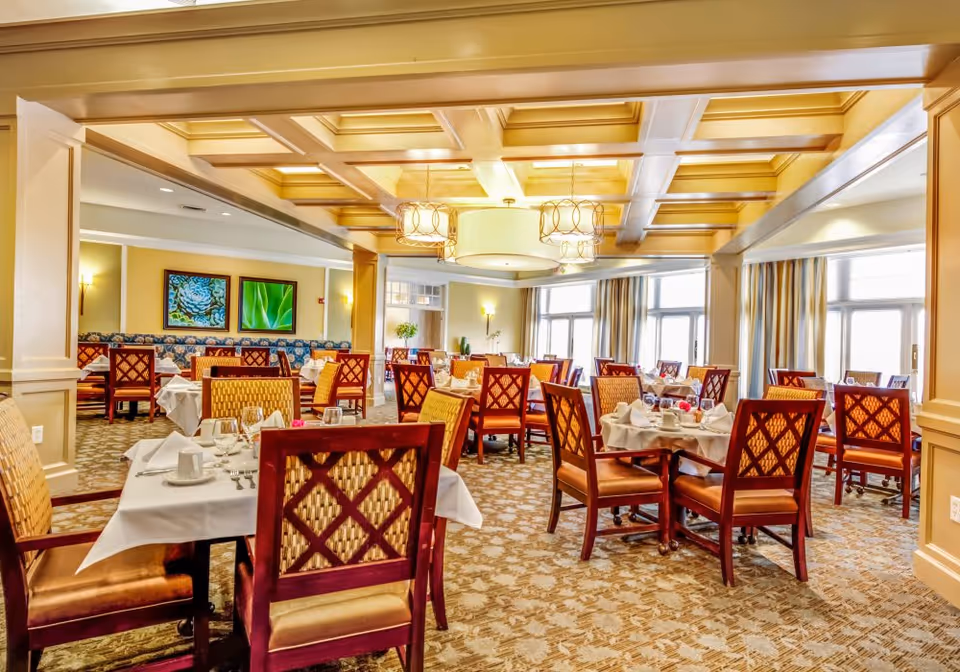 A bright and elegant dining room with multiple tables covered in white tablecloths, set with plates, glasses, and silverware. The room features wooden chairs with patterned upholstery, large windows with curtains allowing natural light, decorative ceiling lights, and framed artwork on the walls.
