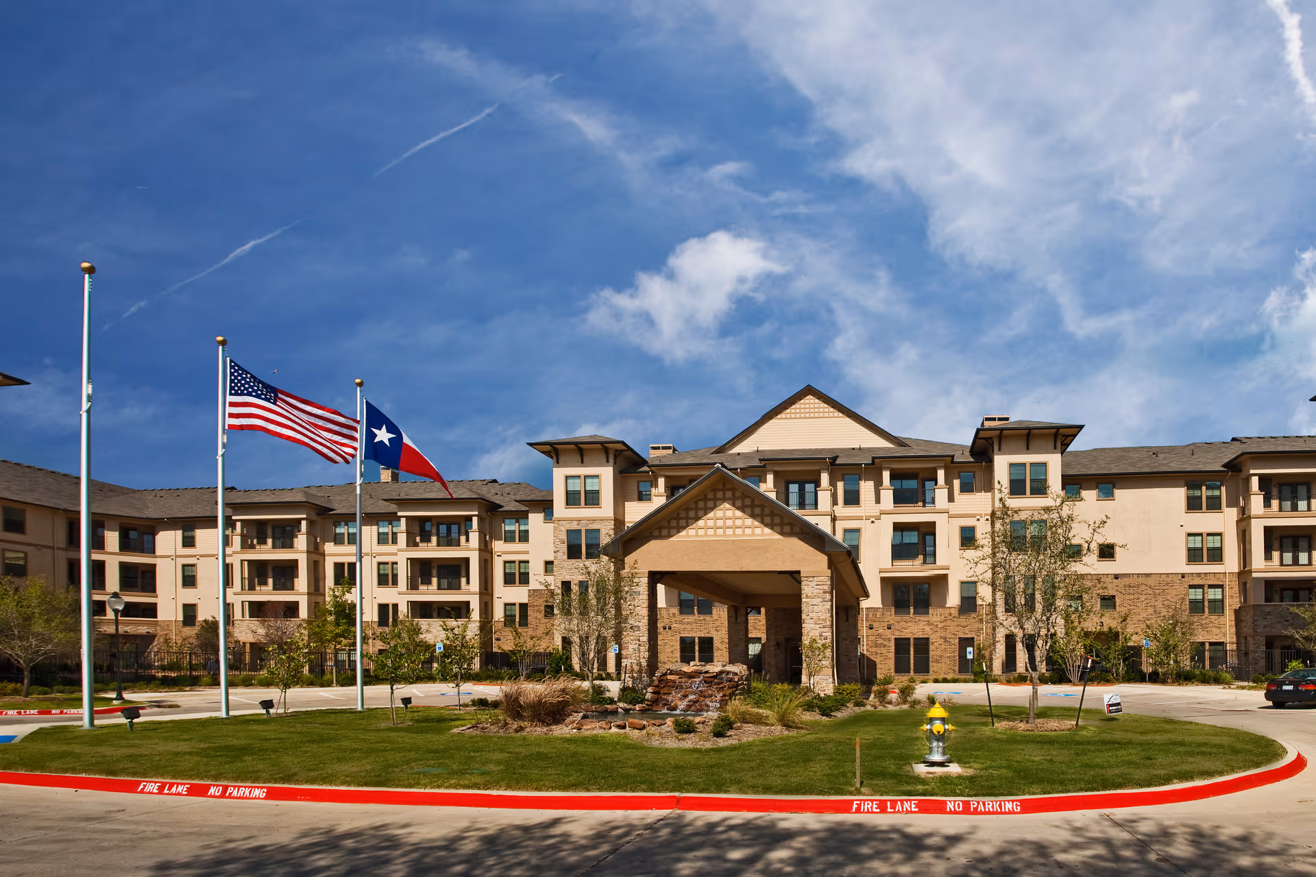 Front exterior view of a large senior living facility building with multiple floors, beige and stone facade, a covered entrance, three flagpoles with American and Texas flags, and a landscaped circular driveway with a fire hydrant and 'Fire Lane No Parking' markings.