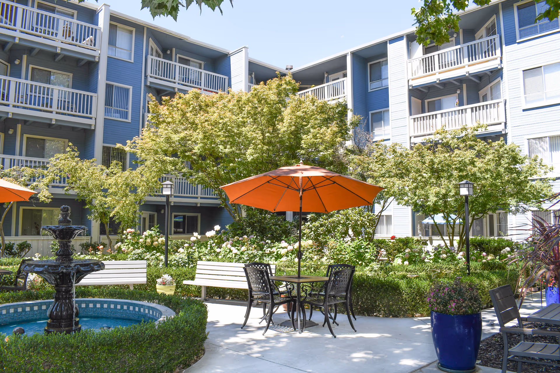Outdoor courtyard area at Carlton Senior Living Concord featuring a black metal table with four chairs under an orange umbrella, surrounded by green bushes, flowering plants, and trees. There is a black tiered fountain on the left side and white benches along the garden path. The background shows a multi-story blue building with balconies and windows.