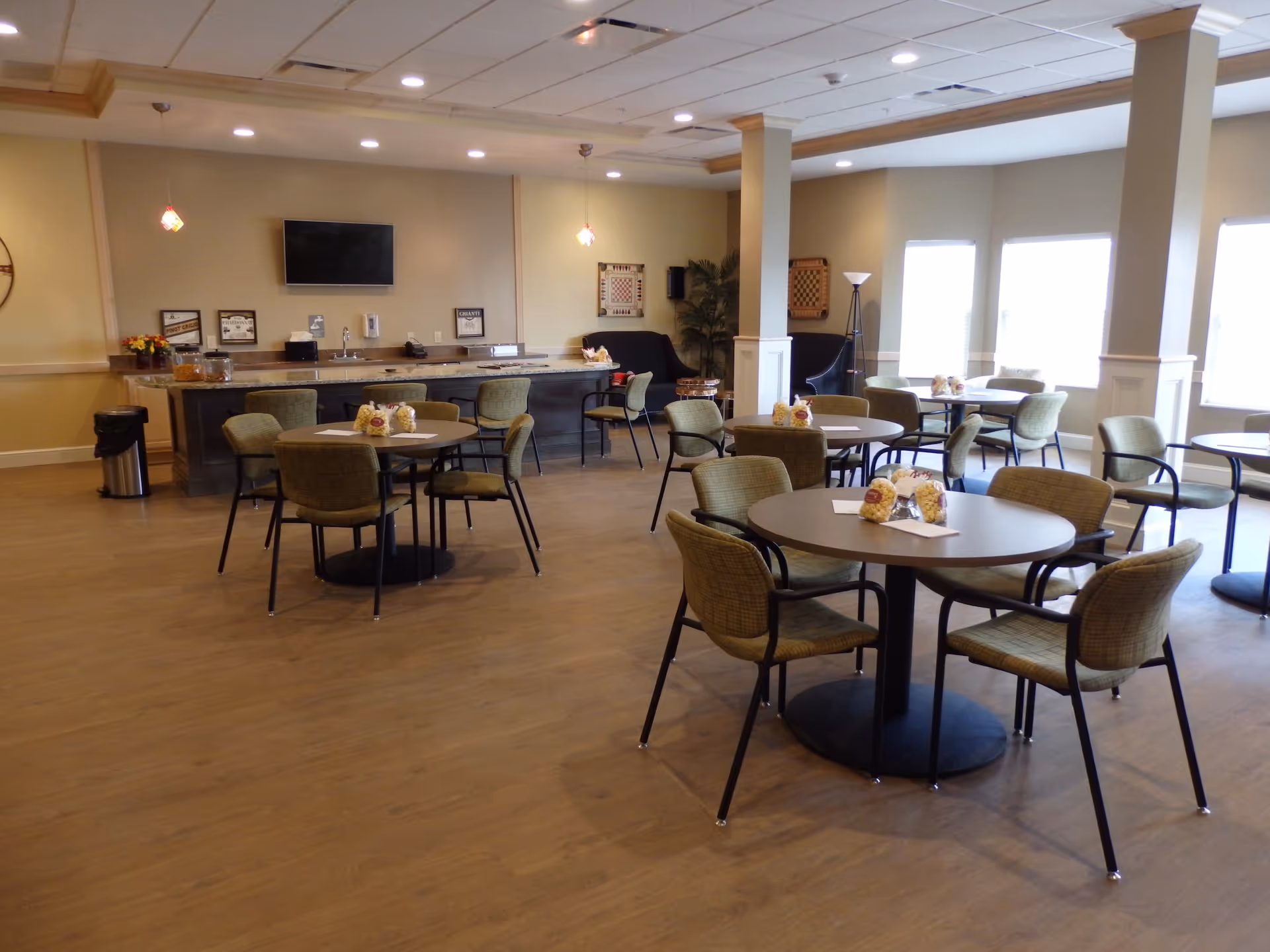 A spacious dining room with several round tables and green cushioned chairs arranged around them. Each table has small bags of popcorn and napkins. The room has large windows letting in natural light, a television mounted on the wall, a counter with a sink, and some decorative items including framed pictures and plants.