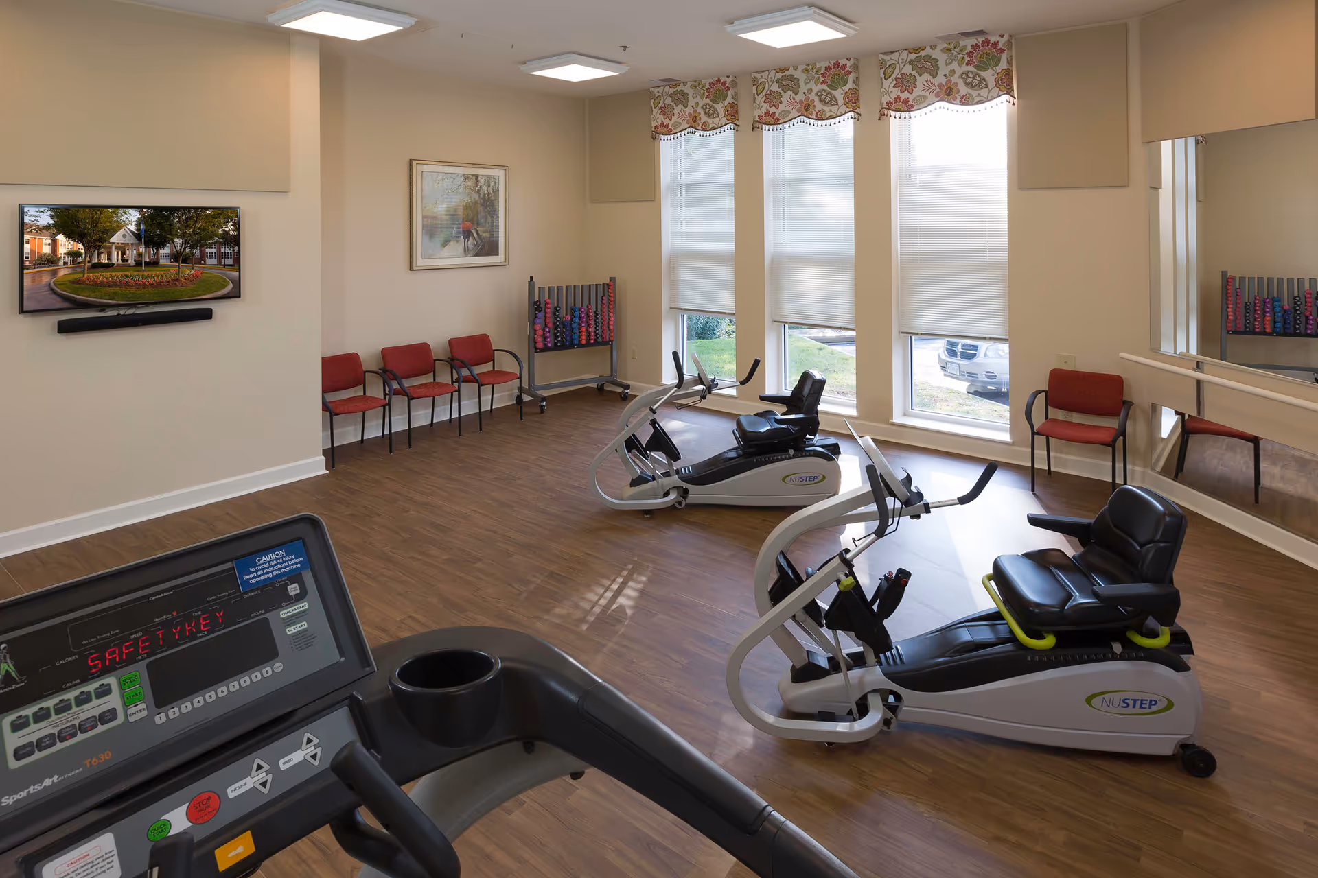 A small fitness room with exercise equipment including a treadmill and two recumbent bikes. The room has wood flooring, three large windows with floral valances and blinds, a wall-mounted TV displaying an outdoor scene, several red chairs along the walls, a rack of colorful dumbbells, and a large mirror on one side.