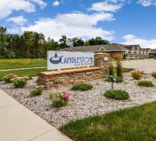 Stone entrance sign reading "Candlestone Assisted Living & Memory Care" in a landscaped gravel bed with flowers and the facility building and driveway behind it under a blue sky.