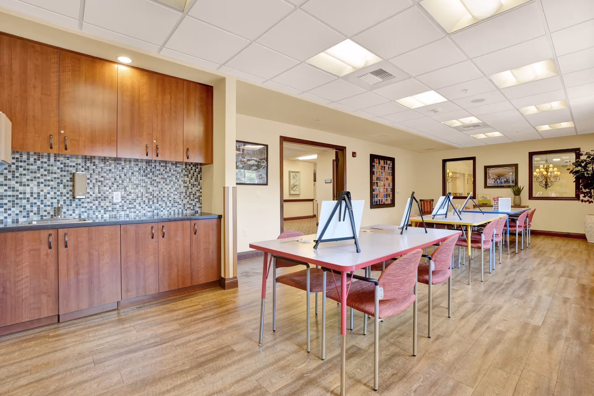 A spacious room with wooden flooring and a kitchenette area featuring wooden cabinets and a mosaic tile backsplash. Several tables with pink chairs are arranged in rows, each table holding easels with blank canvases, suggesting an art or activity room. The room is well-lit with ceiling lights and has framed artwork on the walls.