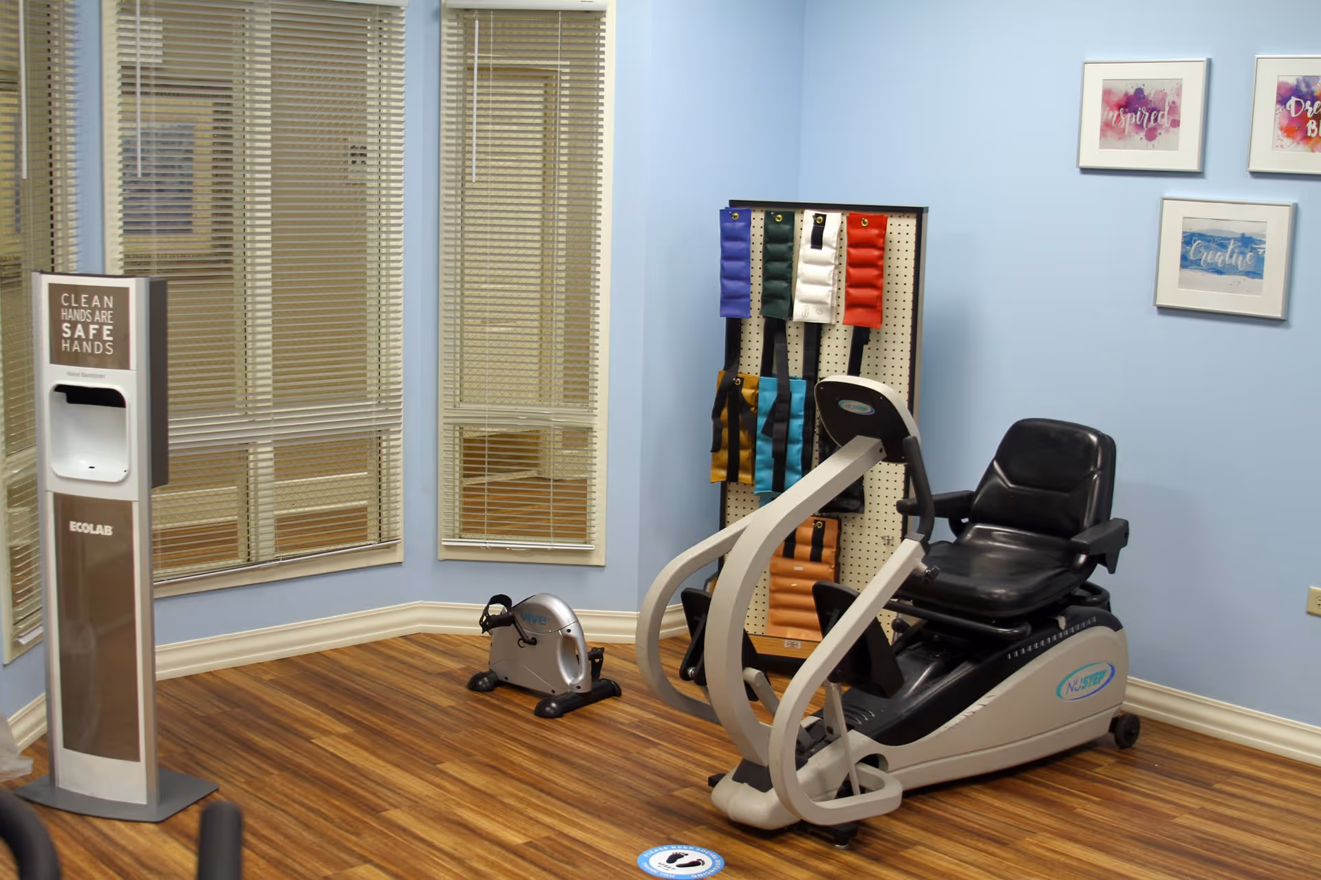 A small exercise room with light blue walls and wood flooring featuring a recumbent exercise bike, a smaller pedal exerciser, a rack with colorful ankle or wrist weights, a hand sanitizer dispenser, and three framed inspirational artworks on the wall.