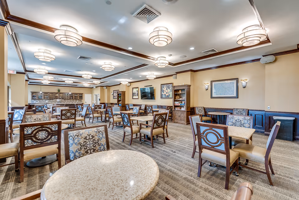 A spacious dining room in a senior living facility with multiple tables and chairs arranged neatly. The room features elegant ceiling lights, framed artwork on the walls, and a carpeted floor. There is a television mounted on the wall and a wooden cabinet with books and decorative items. The overall decor is warm and inviting with beige walls and wooden trim.