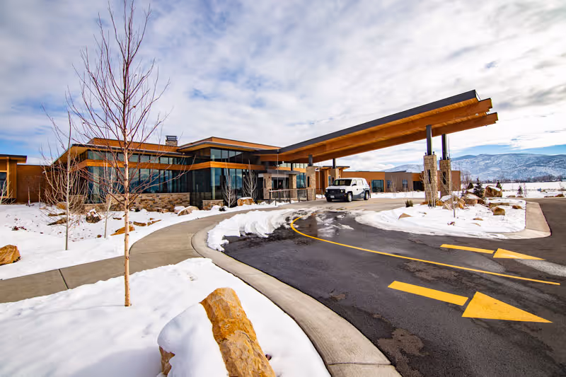 Exterior view of a modern single-story building with large glass windows and wooden architectural accents. The ground is covered with snow, and a curved driveway with yellow directional arrows leads to the entrance where a white vehicle is parked. Leafless trees and rocks are visible in the snowy landscape, with mountains in the background under a partly cloudy sky.