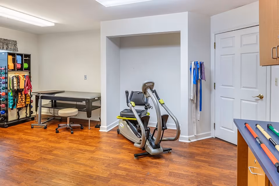 A bright interior therapy/fitness room with a recumbent exercise bike, treatment table, rack of weights and resistance bands on wood floors.