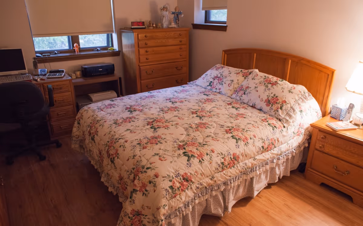 A cozy bedroom with a wooden bed frame and floral-patterned bedding. The room has wooden furniture including a dresser, nightstand with a lamp, and a desk with a computer chair. Two windows provide natural light.