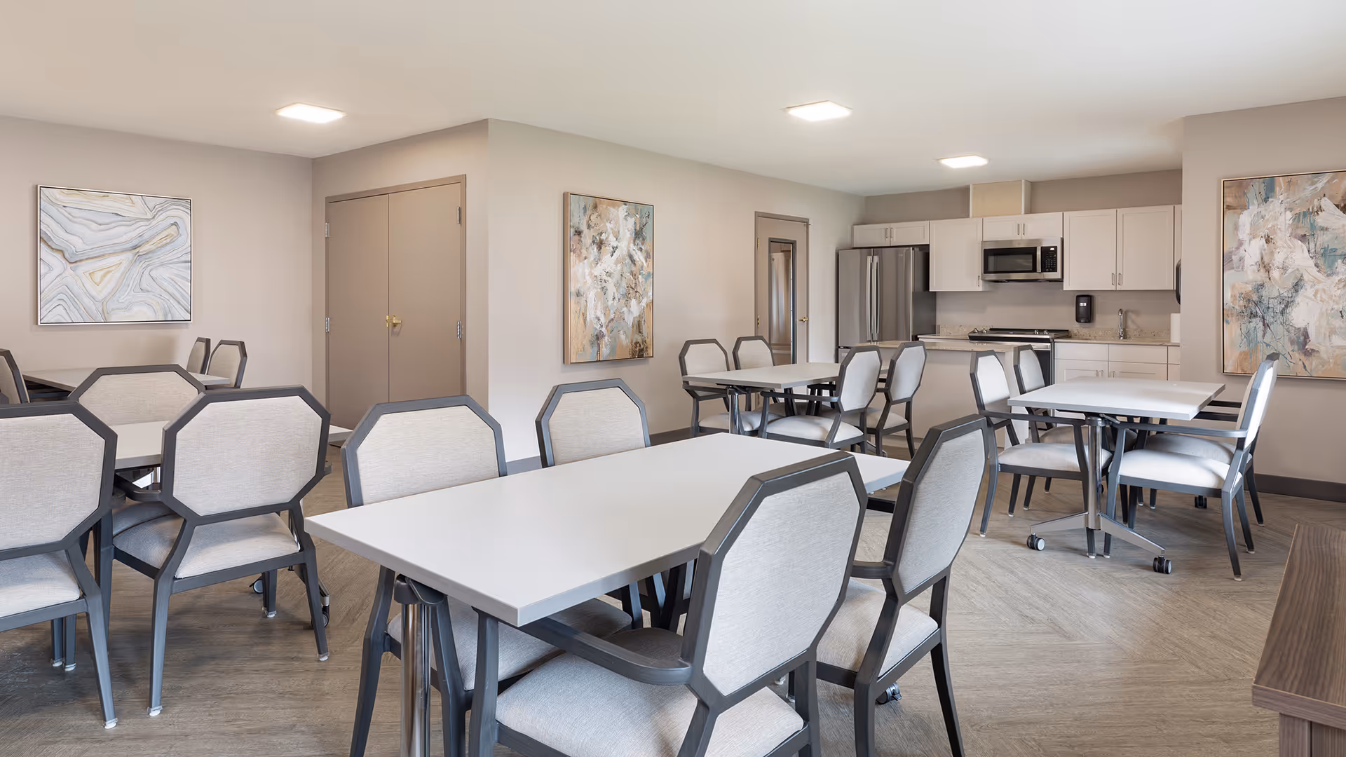 A bright and clean dining room with multiple tables and cushioned chairs arranged neatly. The room features light-colored walls adorned with abstract paintings, a kitchen area with white cabinets, a refrigerator, microwave, and a stove. The flooring is a light wood pattern, and the ceiling has recessed lighting.