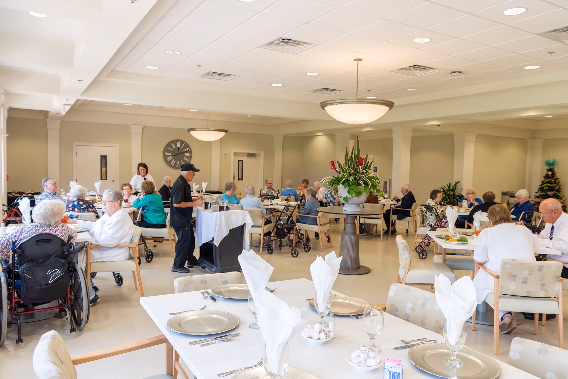 A bright dining room in a senior living facility with elderly residents seated at tables, some in wheelchairs, enjoying a meal. A staff member is serving food from a cart. The room has beige walls, a large clock on the wall, a central table with a large plant, and overhead lighting.