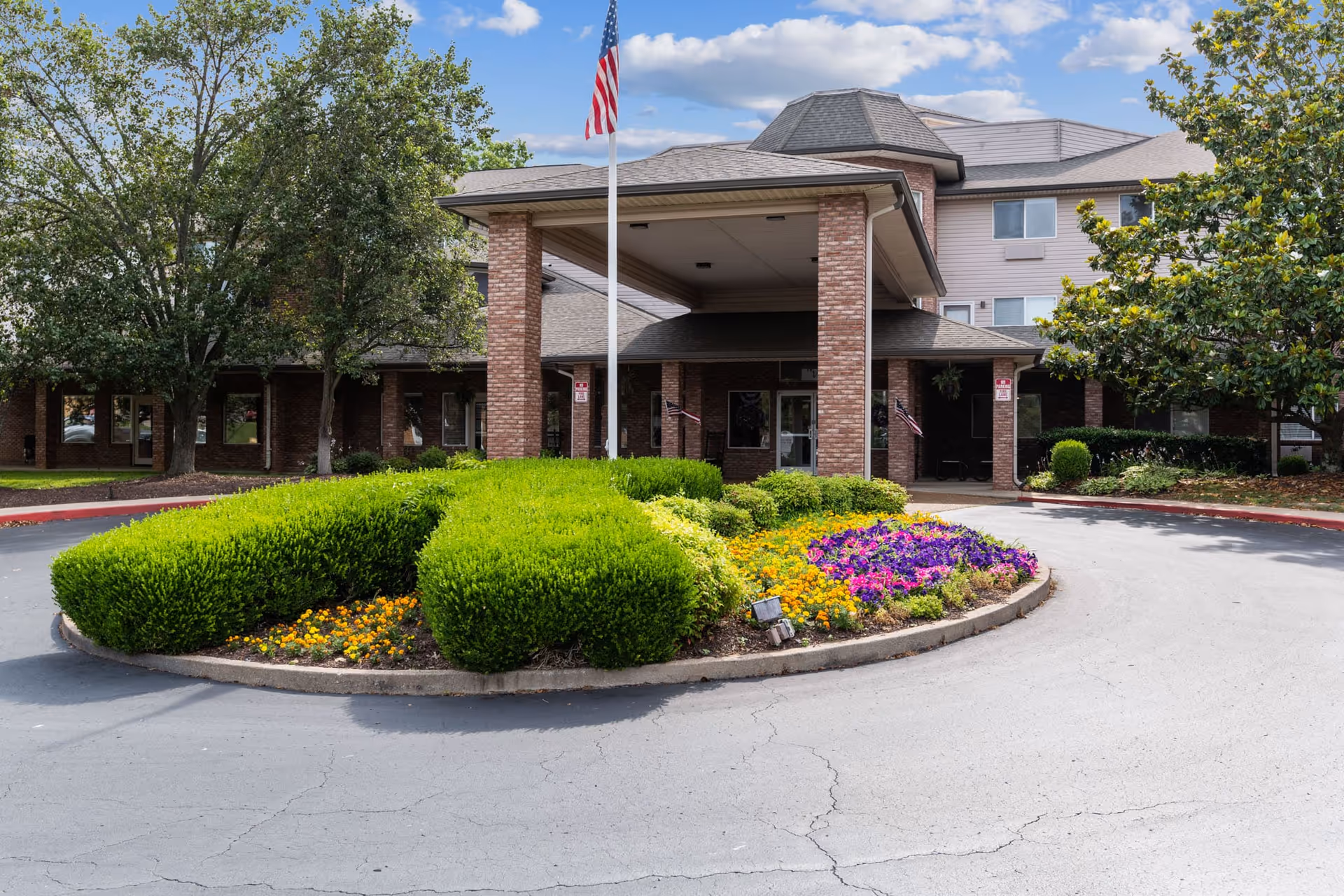 Front entrance of a senior living facility with a covered driveway supported by brick pillars, an American flag on a flagpole, well-maintained green bushes, colorful flower beds, and trees surrounding the building under a partly cloudy sky.
