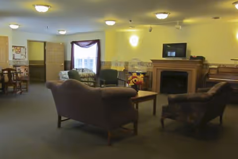 A cozy living room area in an assisted living facility with several upholstered chairs and a sofa arranged around a wooden coffee table with a flower arrangement. There is a fireplace with a TV on the mantel, a piano to the right, and a window with a curtain in the background. The room is softly lit with ceiling lights and wall sconces.