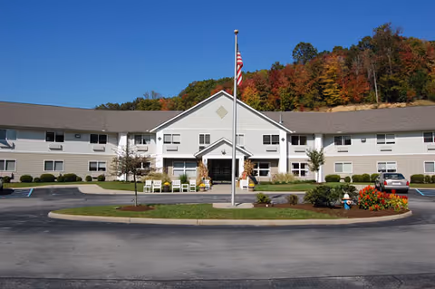 Front exterior view of a two-story senior living facility building with a central entrance, an American flag on a flagpole in front, surrounded by a circular driveway and landscaped greenery, with trees showing fall colors in the background under a clear blue sky.