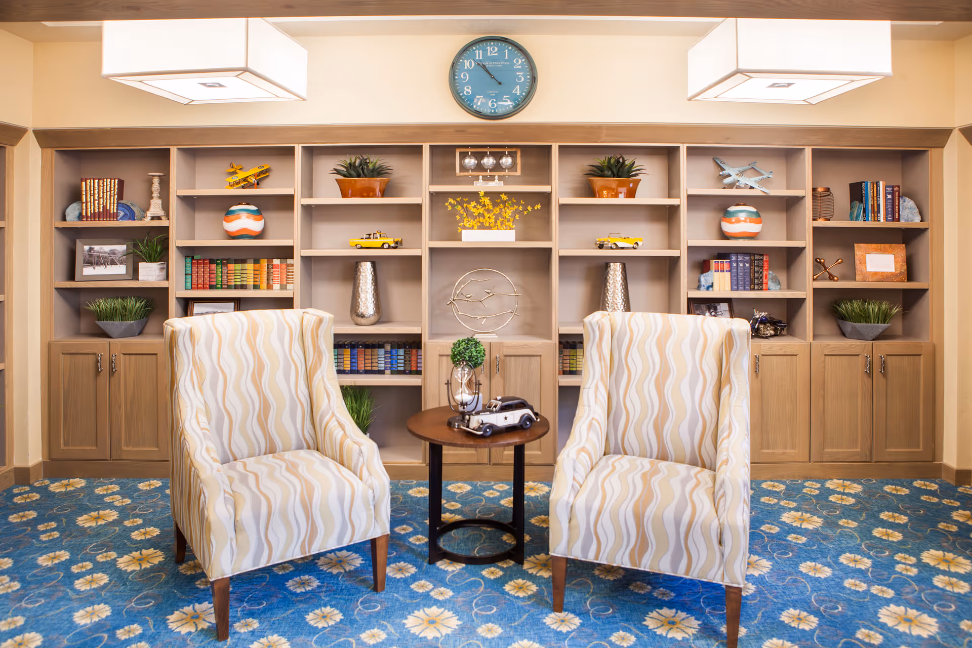 A cozy sitting area with two patterned armchairs facing a small round wooden table. Behind the chairs is a large built-in wooden bookshelf filled with books, decorative items, plants, and model airplanes. A blue clock is mounted on the wall above the bookshelf. The floor is covered with a blue floral carpet, and two rectangular ceiling lights illuminate the space.
