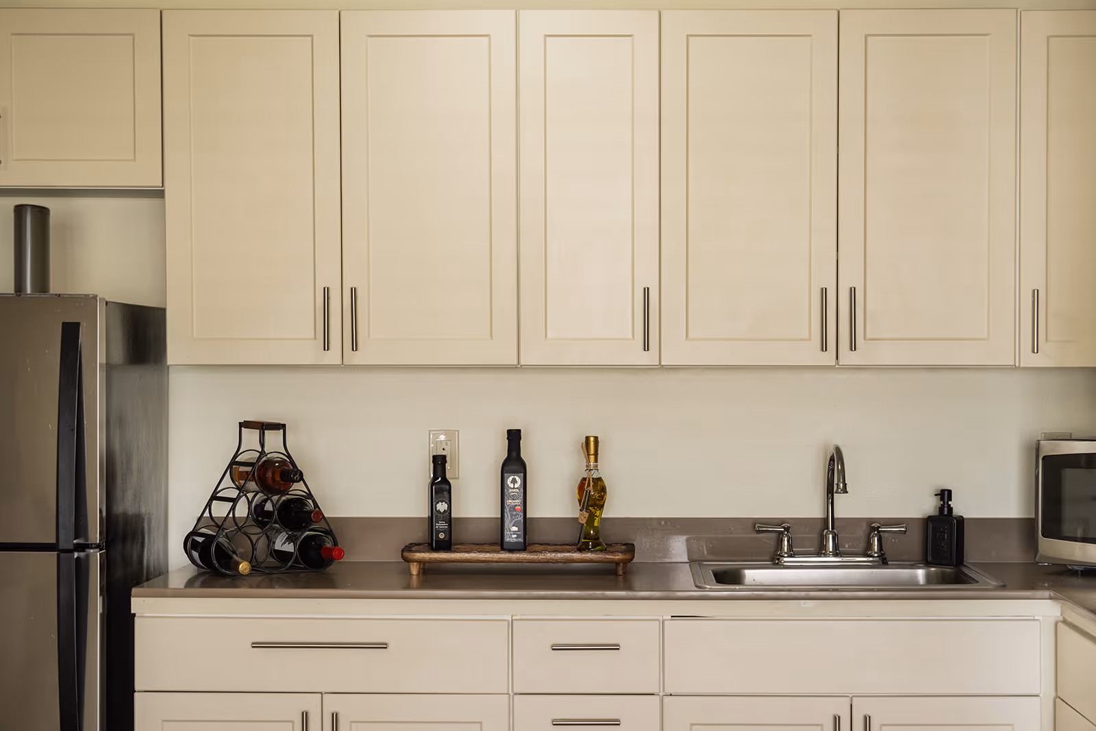 A kitchen countertop with a stainless steel sink and faucet, a microwave on the right, a wine rack with several bottles on the left, and three bottles of oil or vinegar on a wooden tray in the center. Above the countertop are cream-colored cabinets with silver handles.
