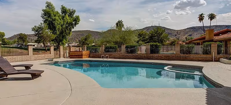 Outdoor swimming pool area with lounge chairs on a concrete deck, surrounded by a low brick wall and metal fence. Trees and hills are visible in the background under a partly cloudy sky.
