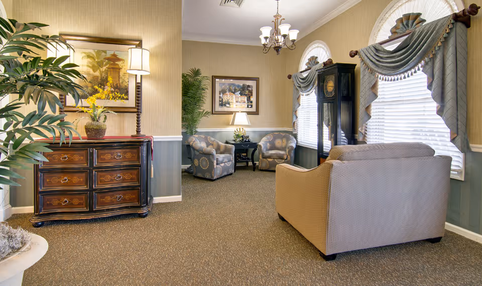 A cozy living room area with beige walls and carpeted floor. The room features a beige sofa, two patterned armchairs, a wooden grandfather clock, and a wooden chest of drawers with a table lamp and a potted plant on top. There are two large windows with decorative valances allowing natural light to enter. The room is decorated with framed artwork and additional plants.