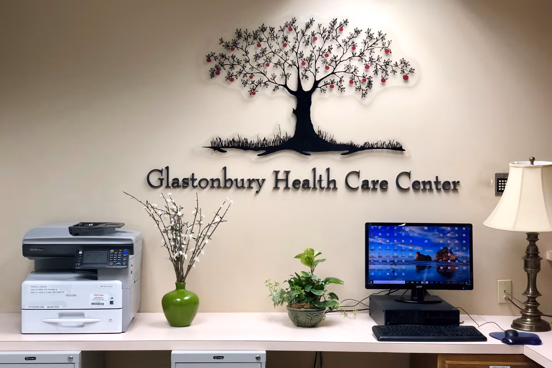 Reception or office area with a printer, two green potted plants, a computer monitor, keyboard, mouse, and a table lamp. On the wall behind is a decorative metal tree with red accents and the text 'Glastonbury Health Care Center'.