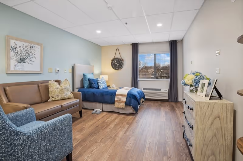 A cozy senior living bedroom featuring a single bed with blue bedding and multiple pillows, a brown leather sofa with a decorative pillow, a blue upholstered armchair, a wooden dresser with framed photos and a vase of flowers, a large wall clock, and a window with dark curtains letting in natural light. The room has wood flooring and light-colored walls.
