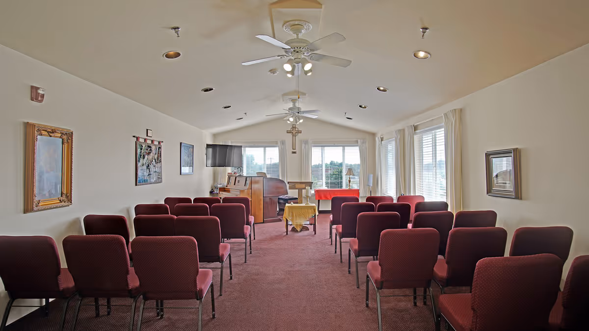 Interior view of a small chapel or meeting room with rows of red cushioned chairs facing a wooden podium and piano. The room has beige walls, several framed pictures, ceiling fans with lights, and large windows with white curtains letting in natural light. A cross is mounted on the wall behind the podium.