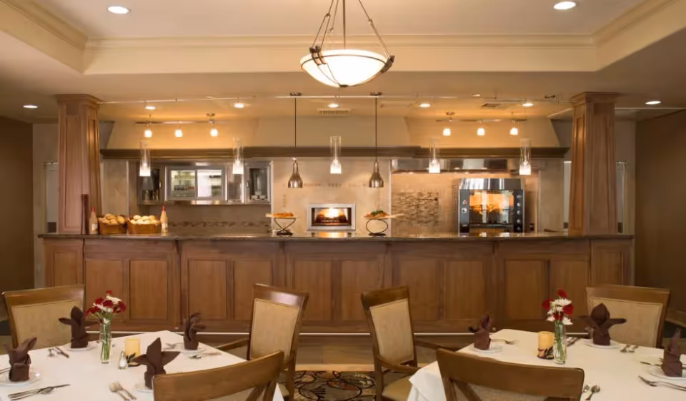 Dining area with tables set with white tablecloths, brown napkins folded in a decorative manner, and small flower vases. In the background, there is a kitchen counter with wooden cabinetry, a bread basket, and modern kitchen appliances including an oven and a microwave. Warm lighting fixtures hang from the ceiling.