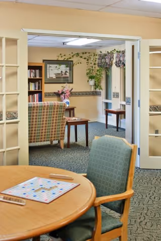 A cozy common area in a senior living facility featuring a round wooden table with a Scrabble game set up, a green cushioned chair, a plaid upholstered armchair, a bookshelf filled with books, framed artwork on the wall, and windows with floral valances and hanging plants.