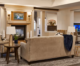 A cozy living room with beige sofas, a side table with a lamp and a plant, a fireplace with a painting above it, and a cabinet with flowers in the background. The room has warm lighting and neutral tones.