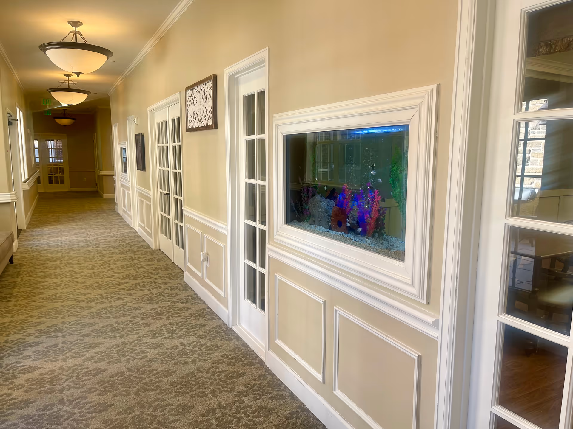 A well-lit hallway in a senior living facility with beige walls and patterned carpet. The hallway features multiple white-framed glass doors and a built-in aquarium with colorful plants and fish. Ceiling lights illuminate the corridor, and there is a bench along the left wall.