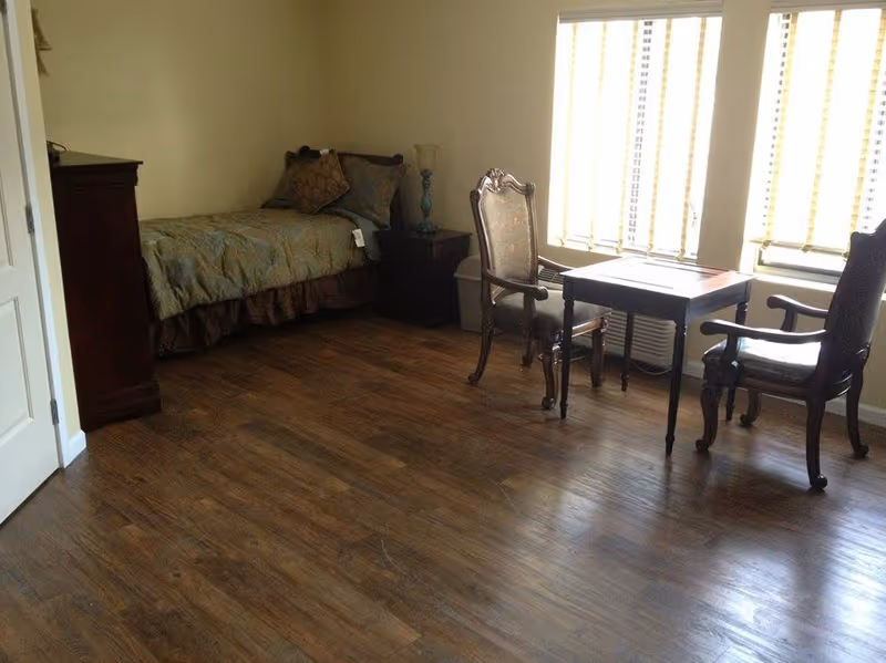 A senior living facility bedroom with a single bed covered in a greenish bedspread and pillows, a wooden nightstand with a lamp, a wooden dresser, and a small table with two ornate wooden chairs near three windows with blinds.