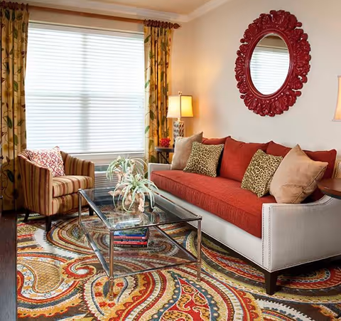 A cozy living room with a red sofa adorned with patterned and solid throw pillows, a striped armchair with a decorative pillow, a glass coffee table with books and a plant, a colorful patterned rug, a large window with floral curtains, a table lamp, and a red decorative round mirror on the wall.