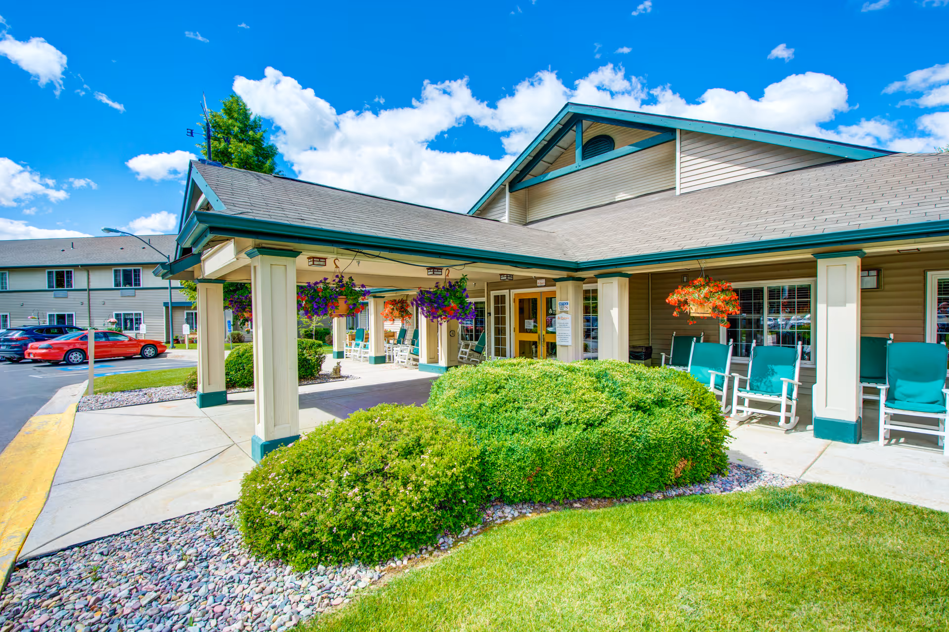 Exterior view of Missoula Valley Senior & Assisted Living facility showing the entrance with a covered porch supported by columns. There are hanging flower baskets, green bushes, and a row of green cushioned rocking chairs along the building. Several cars are parked in the parking lot under a bright blue sky with scattered clouds.