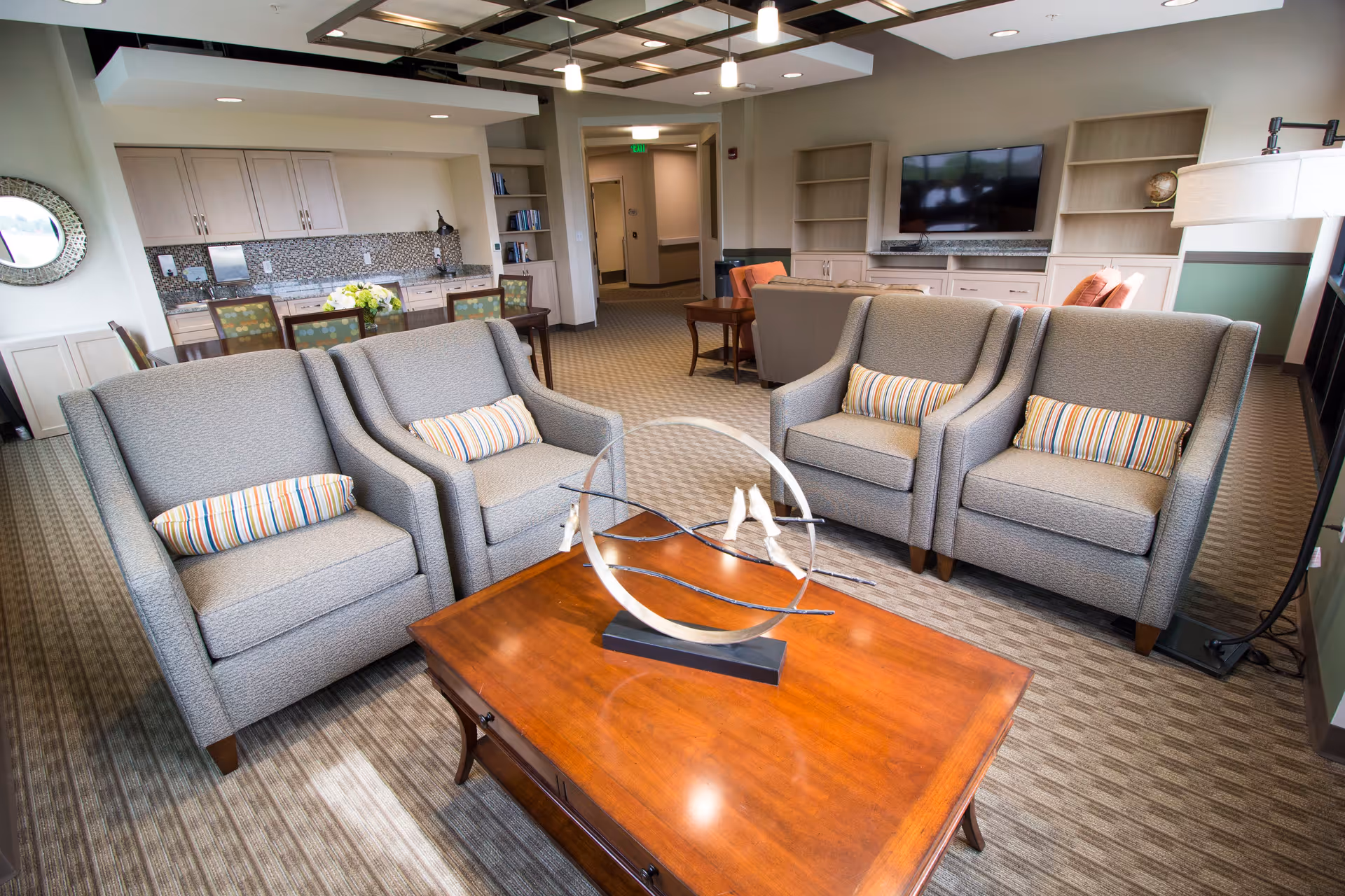 A spacious and well-lit living room area with four gray armchairs arranged around a wooden coffee table featuring a modern decorative sculpture. In the background, there is a flat-screen TV mounted on the wall with built-in shelves and cabinets below. A dining table with chairs is visible near the kitchenette area with cabinets and a mosaic backsplash. The room has a carpeted floor and a ceiling with exposed wooden beams and recessed lighting.