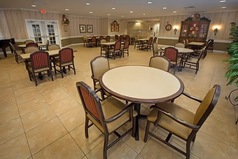 A spacious dining room with multiple round and rectangular tables surrounded by chairs. The room has tiled floors, beige walls with a chair rail, and decorative wall art. There is a wooden cabinet with glass doors on the right side and a potted plant in the corner.