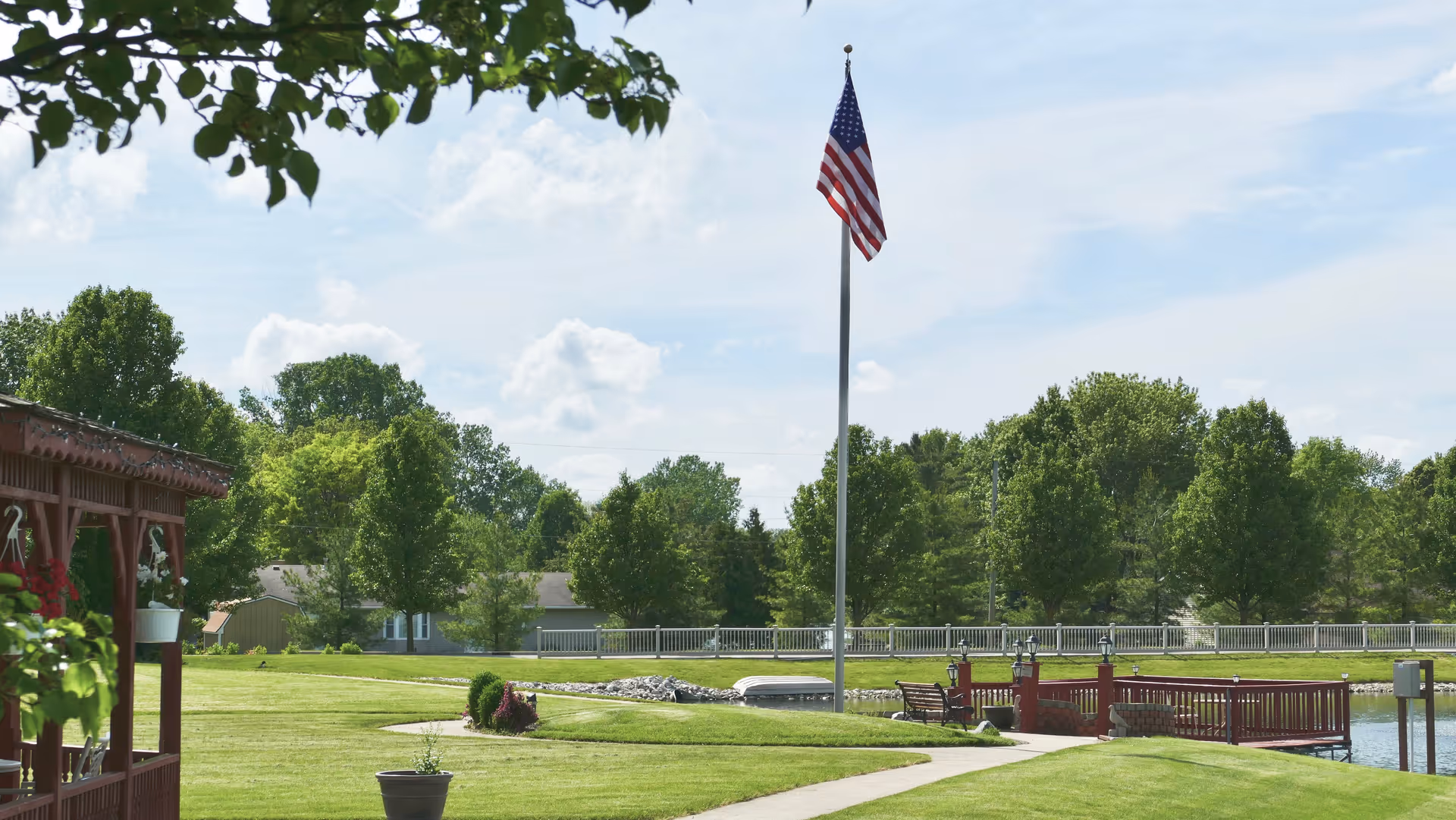 A well-maintained outdoor area featuring a green lawn, a tall flagpole with an American flag, a wooden gazebo with hanging flower pots on the left, a small wooden dock extending over a pond, and trees in the background under a partly cloudy sky.