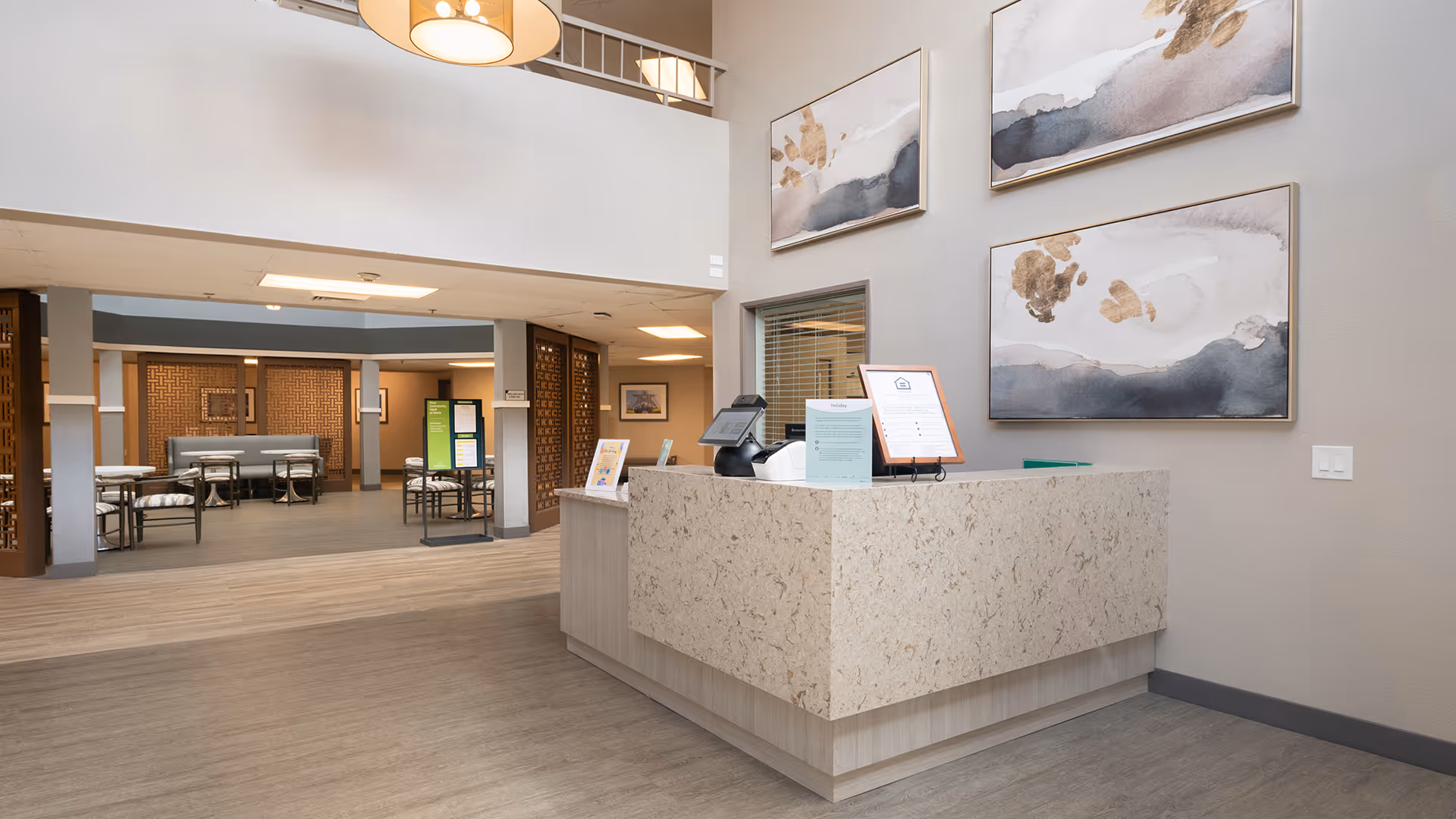 Reception area of a senior living facility with a light-colored stone front desk, decorative abstract paintings on the wall, and a view into a dining area with tables and chairs in the background.